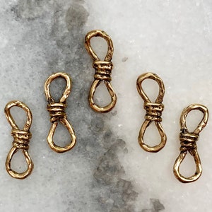 May include: Five gold-toned metal charms shaped like a knot. The charms are on a white marble surface.