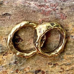 May include: A close-up shot of a gold-colored, double-hoop ring. The ring has a textured, organic design with an irregular shape. The background is a mottled, earthy brown and tan surface.