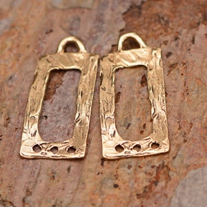 May include: Two gold-colored rectangular pendants with a textured, hammered appearance. Each pendant has a rectangular cutout, a small loop at the top, and three small holes at the bottom. The pendants are displayed on a brown, textured surface.