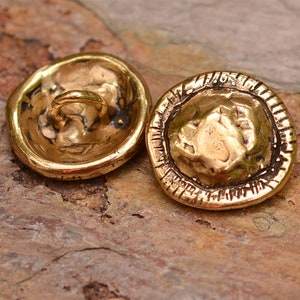 May include: Two gold-toned metal buttons with a textured surface. One button has a raised design and the other has a loop on the back.