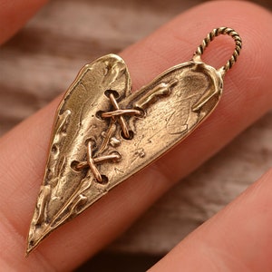 May include: A bronze heart-shaped pendant with a textured surface and a stitched design. The pendant has a twisted loop at the top for hanging. The heart is split and stitched together with cross-shaped accents.