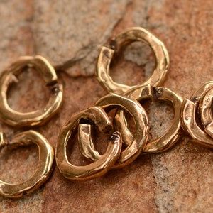 May include: Close-up of several small, gold-colored, hammered metal jump rings. The rings are open and ready to be connected. The background is a textured, brown surface.