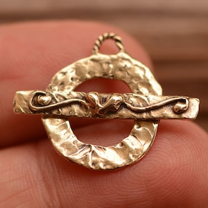 May include: Gold-toned metal clasp with a textured, hammered finish. The clasp features a decorative heart design and a loop for attaching to a chain or cord.