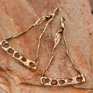 May include: Two gold-colored, abstract triangle-shaped jewelry components. Each component has a series of circular holes along the bottom edge and a loop at the top. The metal has a textured, organic appearance.