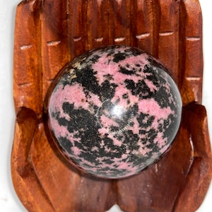 May include: A decorative wooden hand-carved holder cradles a speckled sphere. The sphere features a mix of pink and black colors. The wooden holder is a rich brown color.