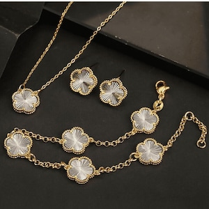 Puede incluir: Un conjunto de joyas de tono dorado con un collar, una pulsera y pendientes. Cada pieza presenta un delicado colgante de trébol transparente con un diseño texturizado.
