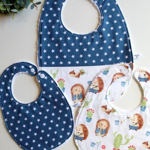 Könnte beinhalten: Drei Baby-Lätzchen, eines mit blauem Hintergrund und weißen Sternen, eines mit weißem Hintergrund und einem Igelmuster und eines mit weißem Hintergrund und einem Igelmuster. Die Lätzchen haben einen weißen Frottee-Saum.