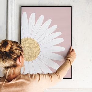May include: A framed print of a large white daisy with a yellow center on a light pink background.