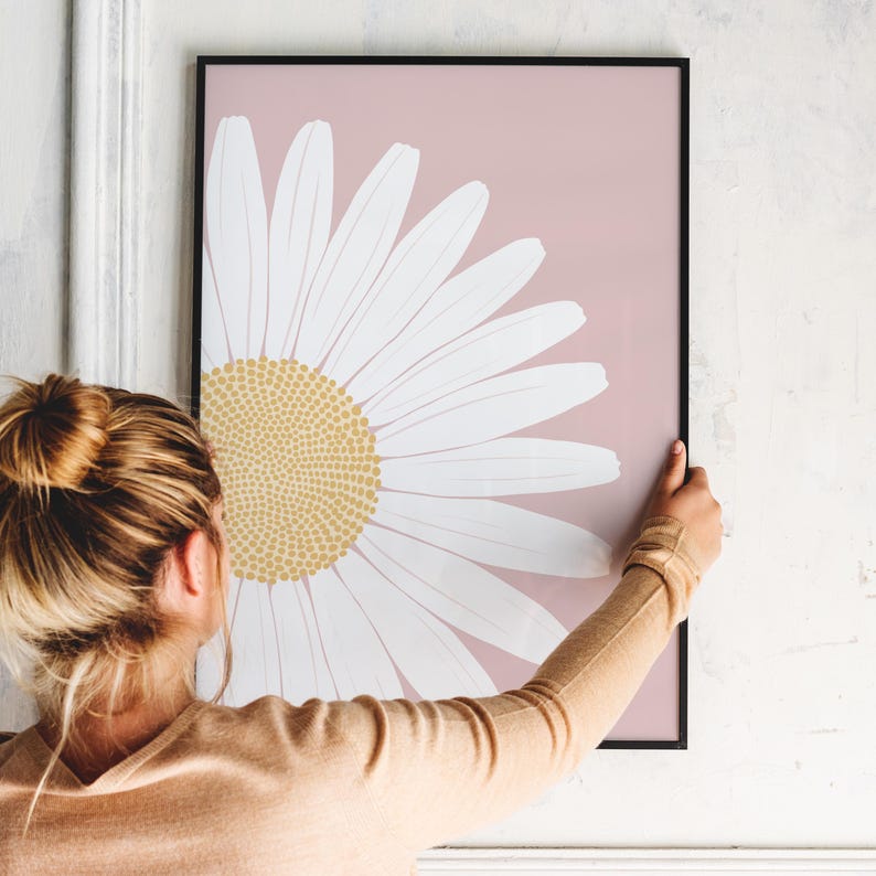 May include: A framed print of a large white daisy with a yellow center on a light pink background.