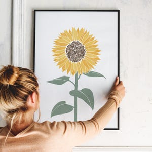 K&ouml;nnte beinhalten: Ein gerahmter Kunstdruck mit einer zentrierten Illustration einer einzelnen Sonnenblume mit leuchtend gelben Bl&uuml;tenbl&auml;ttern und einem dunkelbraunen Zentrum auf wei&szlig;em Hintergrund. Die Sonnenblume hat einen gr&uuml;nen Stiel und vier gr&uuml;ne Bl&auml;tter.