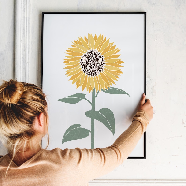K&ouml;nnte beinhalten: Ein gerahmter Kunstdruck mit einer zentrierten Illustration einer einzelnen Sonnenblume mit leuchtend gelben Bl&uuml;tenbl&auml;ttern und einem dunkelbraunen Zentrum auf wei&szlig;em Hintergrund. Die Sonnenblume hat einen gr&uuml;nen Stiel und vier gr&uuml;ne Bl&auml;tter.