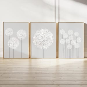 Op de afbeelding: Drie ingelijste kunstprints met witte bloemmotieven op een lichtgrijze achtergrond. De prints hebben goudkleurige frames en worden gepresenteerd op een lichte houten vloer. De bloemmotieven variëren in grootte en opstelling.