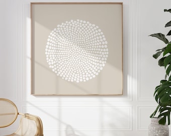 May include: A framed abstract art print featuring a beige background with a white, circular pattern of small dots. The artwork is displayed on a white wall, with a wooden frame. A rattan chair and a potted plant are visible.