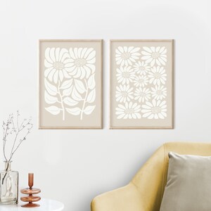 May include: Two framed prints with a beige background and white floral designs. The print on the left features a single large flower with leaves. The print on the right features a repeating pattern of smaller flowers.