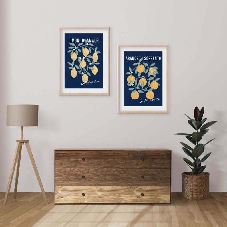May include: Two framed wall art prints featuring botanical illustrations on a dark blue background. The left print displays yellow lemons with the text Limoni di Amalfi and La Dolce Vita. The right print shows orange fruit with Arance di Sorrento and La Vita e Bella.