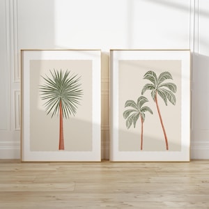 May include: Two framed art prints with palm tree illustrations. One print shows a single palm with green fronds and a red trunk. The other print displays two palms with green fronds and red trunks. The frames are gold, and the background is a neutral beige.