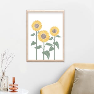K&ouml;nnte beinhalten: Ein gerahmter Wandkunstdruck mit drei Sonnenblumen mit gelben Bl&uuml;tenbl&auml;ttern und braunen Mitten auf wei&szlig;em Hintergrund. Die Sonnenblumen haben lange gr&uuml;ne St&auml;ngel und gr&uuml;ne Bl&auml;tter, alles in einem hellen Holzrahmen eingefasst.