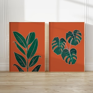 Puede incluir: Dos estampas botánicas enmarcadas. La estampa izquierda presenta una planta verde con hojas grandes sobre un fondo naranja. La estampa derecha muestra un grupo de hojas de monstera verdes sobre fondo naranja. Ambas estampas tienen un marco dorado.