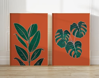 May include: Two framed botanical prints. The left print features a green plant with large leaves against an orange background. The right print shows a cluster of green monstera leaves on an orange background. Both prints have a gold frame.