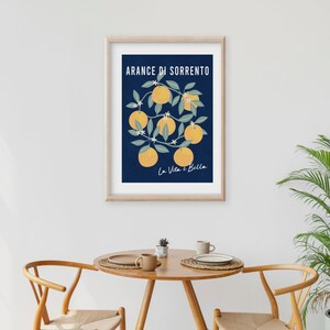 May include: A framed wall art print featuring a dark blue background with a central illustration of yellow oranges on green leafy branches. The text ARANCE DI SORRENTO is printed in white at the top, and La Vita &egrave; Bella is in white script below.