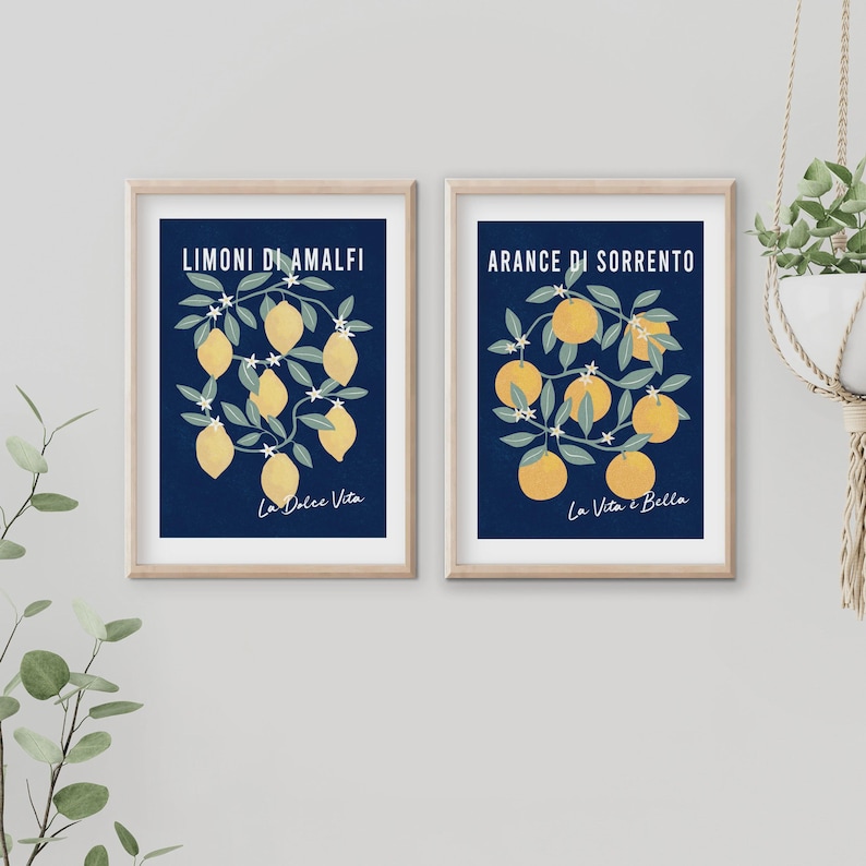 May include: Two framed wall art prints with light wood frames. The left print features yellow lemons on branches with white flowers on a dark blue background, labeled LIMONI DI AMALFI and La Dolce Vita. The right print shows orange oranges with white flowers on a dark blue background, labeled ARANCE DI SORRENTO and La Vita &egrave; Bella.