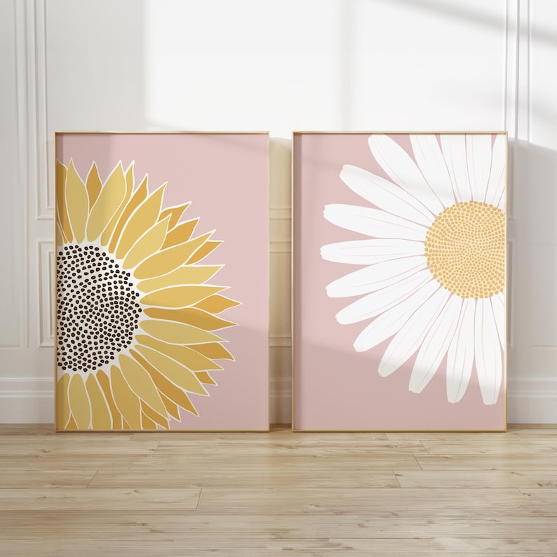May include: Two framed prints with a pink background. The print on the left features a yellow sunflower with a brown center. The print on the right features a white daisy with a yellow center.