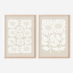 May include: Two beige framed prints with white floral designs. The print on the left features a repeating pattern of stylized white flowers. The print on the right features a cluster of stylized white flowers with leaves.