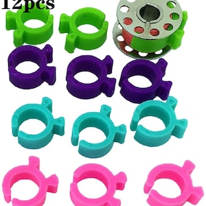 May include: Twelve colorful plastic thread bobbin holders in green, purple, turquoise, and pink. The bobbin holders are designed to hold thread spools for sewing machines. The image also includes a metal bobbin winder with a red and green thread spool.
