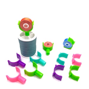May include: A set of colorful thread bobbins for sewing. The bobbins are made of plastic and have a U-shaped design. There are 9 bobbins in total, including pink, purple, green, blue, and orange. The bobbins are arranged on a white surface.
