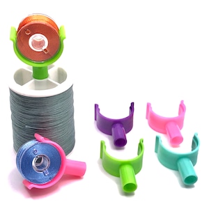 May include: A set of four colorful thread bobbins with a spool of gray thread and a bobbin holder. The bobbins are purple, pink, green, and blue.