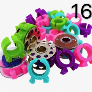 May include: A set of 16 colorful thread bobbin holders for sewing machines. The holders are made of plastic and come in a variety of colors, including green, pink, purple, and blue. The holders are designed to fit over the bobbin and help keep the thread from tangling.