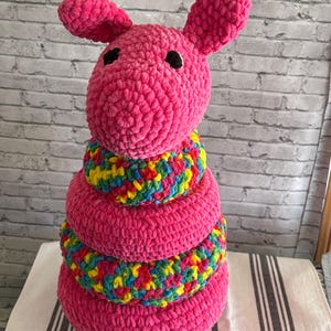 Puede incluir: Un juguete apilable de conejito rosa tejido a mano con un anillo de colores arcoíris. El conejito tiene ojos negros y orejas grandes. El juguete está hecho de hilo suave y afelpado y está diseñado para bebés y niños pequeños.