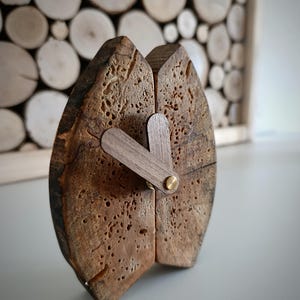 Könnte beinhalten: Eine einzigartige Holzuhr mit natürlicher, strukturierter Optik. Das Zifferblatt besteht aus zwei Holzstücken mit dunkelbraunem Finish und sichtbarer Maserung. Die Zeiger sind hellbraun, vor einem Hintergrund aus Holzscheiben.