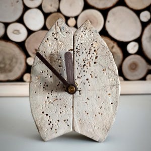 Könnte beinhalten: Eine handgefertigte Holzuhr im rustikalen und natürlichen Design. Das Zifferblatt besteht aus zwei Teilen hellfarbenem, strukturiertem Holz. Sie hat dunkelbraune Zeiger und einen zentralen Holzpfosten. Das Design hebt die Holzmaserung und kleine Löcher hervor.