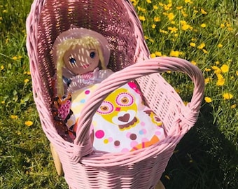 Puede incluir: Un cochecito de muñecas de mimbre rosa con una muñeca dentro. La muñeca lleva un vestido blanco y un sombrero. El cochecito tiene un forro de estampado de búho blanco y rosa.