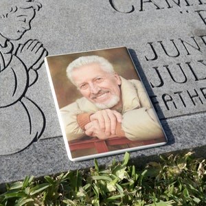 May include: A black and white drawing of a praying angel is carved into a gray granite headstone. A white ceramic tile with a photo of a smiling man is placed on the headstone. The tile reads "June July Father".