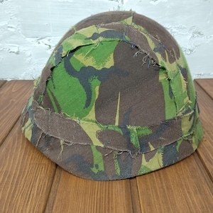 Puede incluir: Un casco con patrón de camuflaje en tonos verdes, marrones y negros. El casco tiene forma redondeada y está hecho de tela. Los bordes tienen un aspecto deshilachado. El diseño de camuflaje es clave.