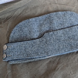 Könnte beinhalten: Eine graue Wollmütze im Militärstil mit schwarzem Band und zwei silbernen Knöpfen mit Kreuzdesign.