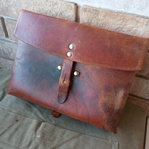 May include: A brown leather satchel with a flap closure and a leather strap secured by two metal rivets. The satchel has a worn appearance, with darker areas and a visible texture. The bag is resting on a green fabric.
