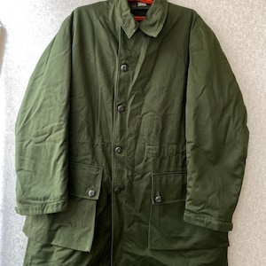 Puede incluir: Abrigo verde oliva con botones, cuello y múltiples bolsillos. El abrigo está colgado de una percha roja. El abrigo parece estar hecho de un material duradero, posiblemente resistente al agua.