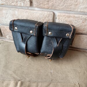 Puede incluir: Un par de bolsas de cuero negro texturizado con una forma rectangular estructurada. Cada bolsa cuenta con una solapa asegurada por un remache de metal plateado y una correa de cuero vertical. Los bordes están acabados con costuras en color canela.