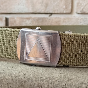 May include: An olive green canvas belt featuring a rectangular silver-toned metal buckle. The buckle displays a centered engraved triangle surrounded by horizontal line detailing. The belt strap is made of a woven fabric material with a textured finish.
