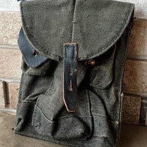Könnte beinhalten: Eine olivgrüne Canvas-Tasche im Vintage-Stil mit mehreren Taschen und einem Klappverschluss. Die Tasche hat schwarze Lederdetails an der Klappe und am Riemen, die mit einem Messingknopf befestigt sind. Das Canvas weist Gebrauchsspuren auf, was ihm ein robustes, gealtertes Aussehen verleiht.