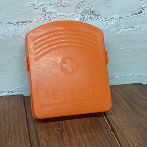 May include: An orange first aid kit with a white cross symbol and the word "АПТЕЧКА" printed on the front. The kit is rectangular with rounded edges and a handle. The background is a white textured wall and a wooden surface.
