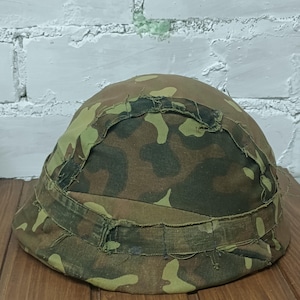 May include: A camouflage-patterned helmet in shades of brown, green, and tan. The helmet has a rounded shape with a fabric band around the base. The fabric shows signs of wear and tear, with frayed edges.