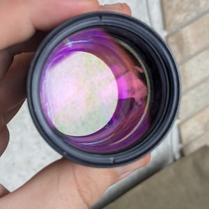 May include: A close-up view of a camera lens, held in a hand. The lens has a black frame and a reflective, multi-colored interior with shades of purple and yellow. The lens is circular and appears to be made of glass.
