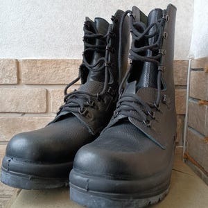 May include: A pair of black leather combat boots with high tops and thick soles. The boots have multiple eyelets for the laces and are designed for rugged use. The boots are displayed on a neutral-colored surface.