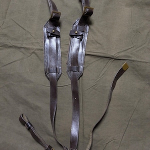 May include: A pair of brown leather straps with brass buckles. The straps are connected at the bottom and have two separate loops at the top.