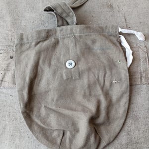 Op de afbeelding: Een beige canvas buidel met een enkele witte knoopsluiting aan de voorzijde, een stoffen lus met knoopsgat aan de bovenkant en een wit trekkoord aan de zijkant. De stof heeft een zichtbare geweven textuur en een afgeronde onderkant.
