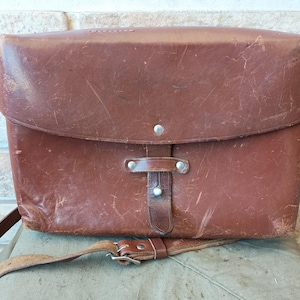 Könnte beinhalten: Eine Vintage-Umhängetasche aus braunem Leder mit Klappenverschluss und verstellbarem Schulterriemen. Die Tasche hat einen Schnallenverschluss und Metallnieten. Das Leder weist Gebrauchsspuren auf, was zu seiner antiken Ästhetik beiträgt.